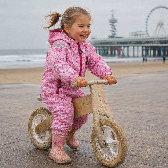 Lachend meisje op een houten loopfiets op het strand, draagt een roze regenpak met bladprint.