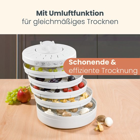 Witte Clatronic voedseldroger met vijf niveaus, gevuld met fruit en paddenstoelen. Naast de droger een tros groente druiven en een rode appel.