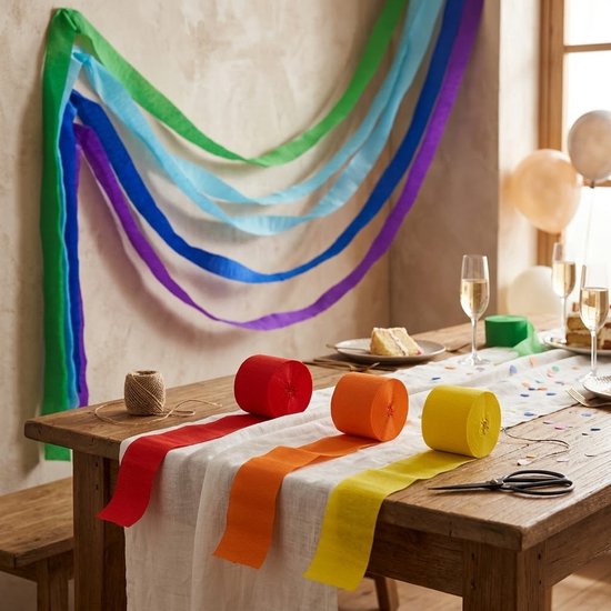 Veelkleurige slingers (regenboog) hangen over een tafel met daarop servies, gebak en rolletjes crêpepapier.