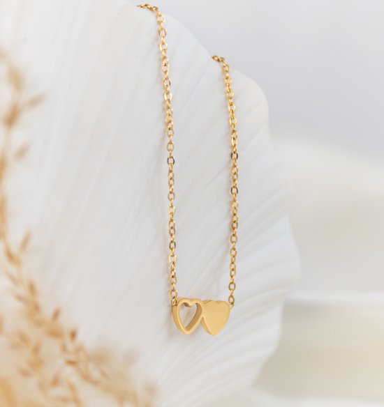 Gouden ketting met twee in elkaar grijpende hartjes aan een dunne schakelketting, gepresenteerd op een witte, schelpachtige achtergrond.