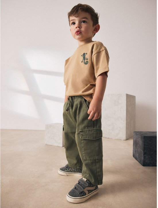 Jongen in een beige T-shirt met krokodillenprint en een groene cargobroek met wijde pijpen.