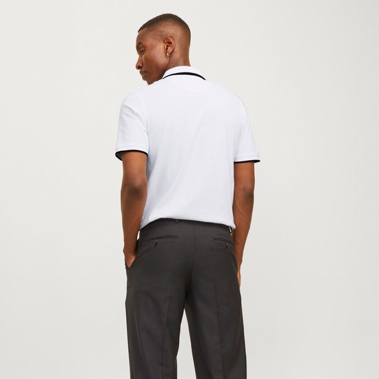 Model draagt wit poloshirt met zwarte biezen, kijkend weg. Zwarte broek.
