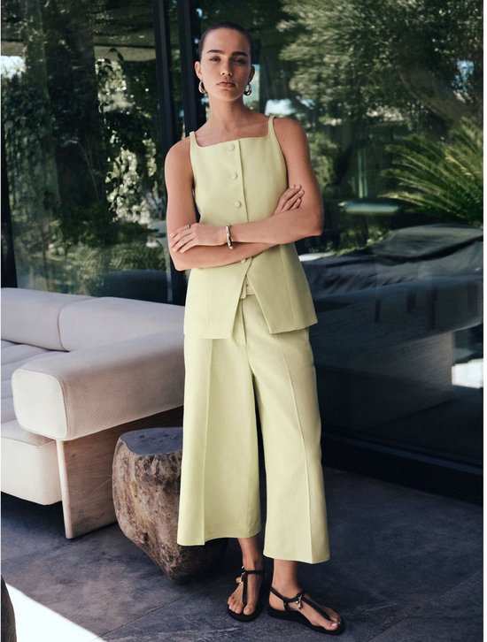 Model draagt een lichtgroene culotte met vierkante hals top, met de armen over elkaar geslagen.