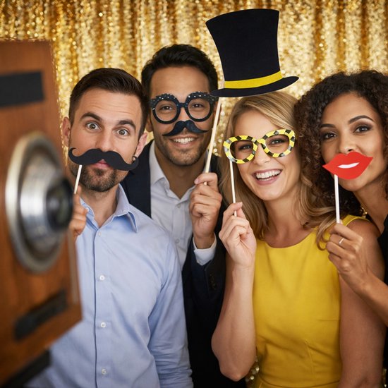 Vier vrienden met fotohokjes-accessoires: man met snor, man met gekke bril, vrouw met gele bril, vrouw met rode lippen en man met hoge hoed.