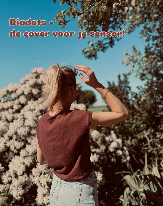 Vrouw met blond staartje, draagt een bruin shirt en een sensor met een bordeauxrode cover op haar bovenarm, in een tuin met bloeiende struiken.