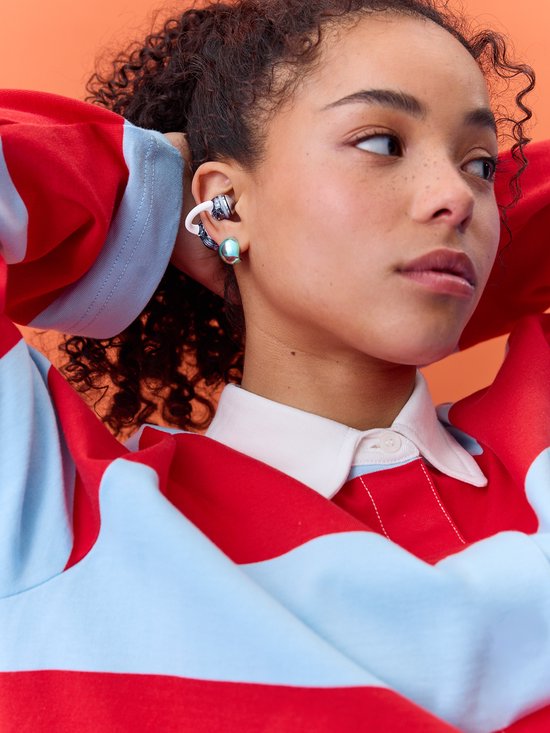 Een vrouw met krullend haar draagt een rood-blauwe gestreepte trui met een witte kraag, met een JBL Soundgear Clip in haar oor.