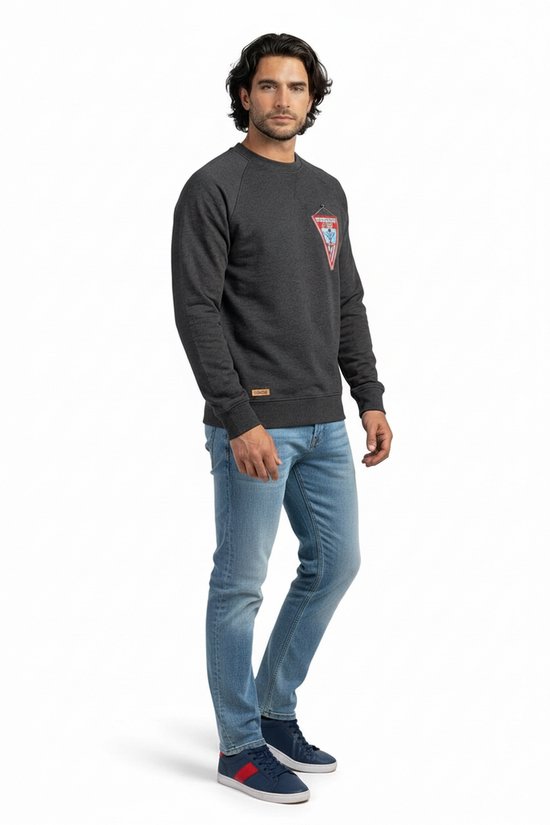Homme portant un pull foncé avec un écusson appliqué sur la poitrine. Jean clair et baskets sombres.