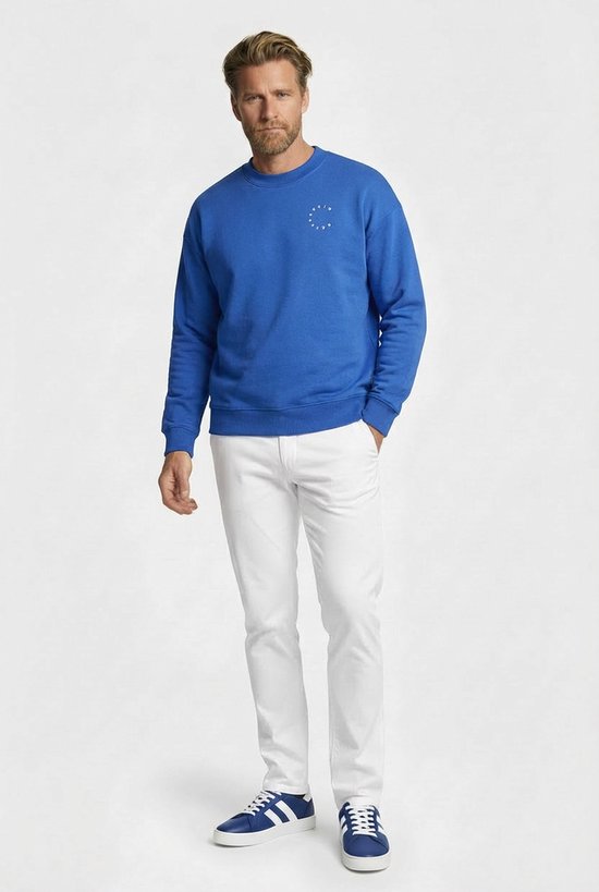 Man met blauwe sweater, witte broek en blauwe sneakers, staand, met witte tekst in cirkel op borst.