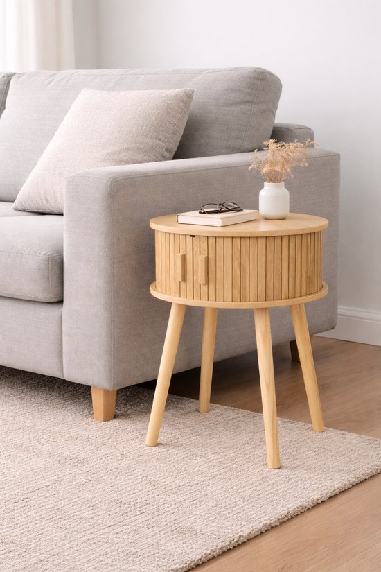 Ronde houten bijzettafel met geribbelde zijkanten en twee deurtjes, naast een grijze bank. Op de tafel liggen een boek en een bril.