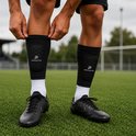 Een persoon trekt zwarte scheenbeschermer sleeves met pads aan over witte sportsokken en zwarte voetbalschoenen.