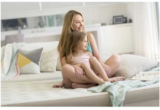 Mère et fille assises et souriantes sur un matelas pneumatique de couleur crème dans une chambre. Elles portent des vêtements confortables.