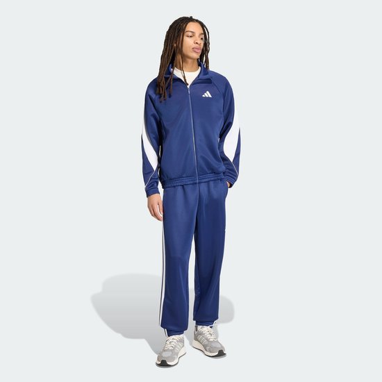 Man met dreadlocks draagt een blauw Adidas trainingspak met witte strepen op de broek.