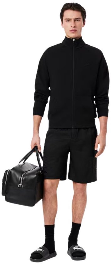 Man met zwarte Lacoste slippers en zwarte kleding, houdt leren tas vast.