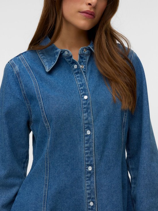Model draagt een denim jurk met lange mouwen in een middellange blauwe denimwas. De jurk heeft een kraag en knoopsluiting aan de voorkant.