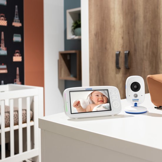 Witte babyfoon met camera, monitor toont baby met handdoek. Op een wit bureau, met kinderledikant op achtergrond.