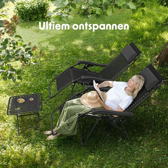 Een vrouw met hoed leest op een zwarte ligstoel in een schaduwrijke tuin. Naast haar staat een bijzettafel met drankjes.