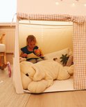 Meisje leest in beige speeltent met bruin-wit geblokte gordijnen en veel knuffels.