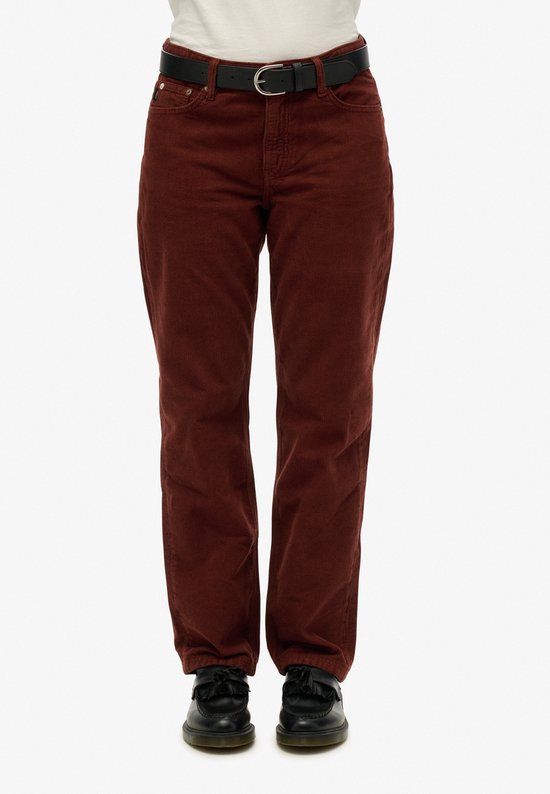 Vrouw in bruine corduroy broek, voorkant zichtbaar. Zwarte riem, witte T-shirt, zwarte schoenen.