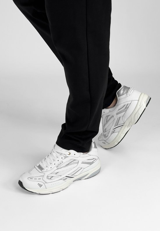 Voeten in witte "DUTCH'D" sneakers met zilveren details, staand op de grond.