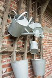 Zinken gieters en emmers hangend aan een houten rek voor een bakstenen muur.