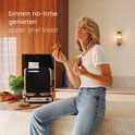 Vrouw met een tosti in elke hand, genietend. Ze zit op het aanrecht, naast een Safecourt contactgrill.
