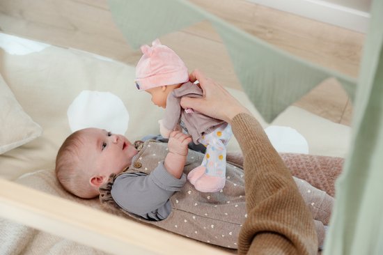 Baby in een beige tuinbroek met stippen en een grijs shirt. Een hand houdt een popje met roze muts en grijs shirt omhoog.