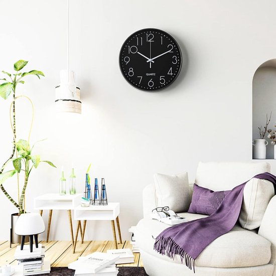 Moderne zwarte wandklok aan een witte muur, boven een bank met paarse deken.