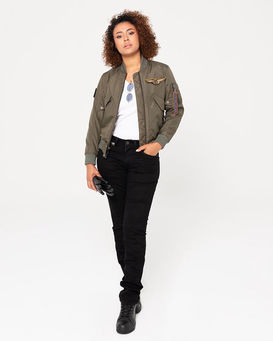 Vrouw in een olijfgroene bomberjack met badge en witte top, zwarte jeans en zwarte schoenen, met zwarte handschoenen in hand.