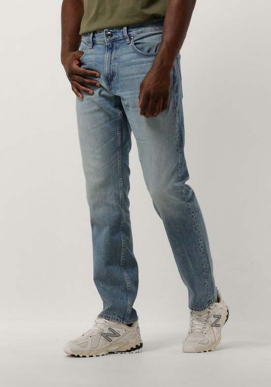 Man met donkere huid draagt een olijfgroene T-shirt met een lichte spijkerbroek en witte sneakers.