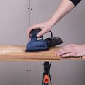 Blauwe FERM schuurmachine met stofzak, in handen van een gebruiker op een houten plank.