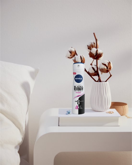 NIVEA Black & White Invisible Original deodorant spray op een nachtkastje met katoenbloemen.