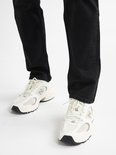 Voeten van een persoon die witte sneakers met mesh details en zwarte jeans draagt.