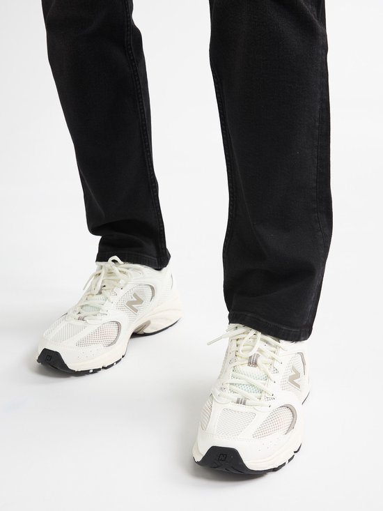 Voeten van een persoon die witte sneakers met mesh details en zwarte jeans draagt.