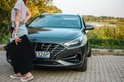 Vrouw met gestreepte top en jeans, houdt zwarte vloermat voor donkergrijze auto.