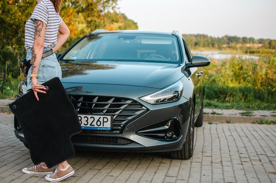 Vrouw met gestreepte top en jeans, houdt zwarte vloermat voor donkergrijze auto.