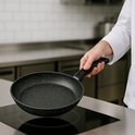Hand houdt een zwarte koekenpan met granieten look boven een inductiekookplaat.