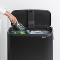 Gros plan d'une poubelle Brabantia noire. Une main laisse tomber une bouteille en plastique dans le compartiment gauche.