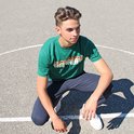 Jonge man zit op basketbalveld, draagt groene t-shirt met tekst en donkere joggingbroek.