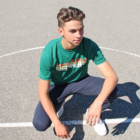 Jonge man zit op basketbalveld, draagt groene t-shirt met tekst en donkere joggingbroek.