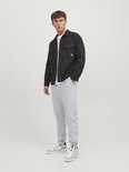 Man die staat met een lichtgrijze joggingbroek, wit T-shirt en een zwart jasje.