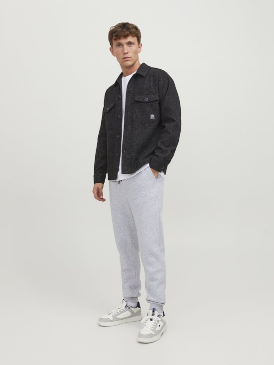 Man die staat met een lichtgrijze joggingbroek, wit T-shirt en een zwart jasje.