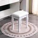 Witte gestoffeerde kruk op een ronde, crèmekleurige vloerkleed met zwarte cirkelvormige patronen. De kruk staat in een slaapkamer met een witte kast en plant op de achtergrond.