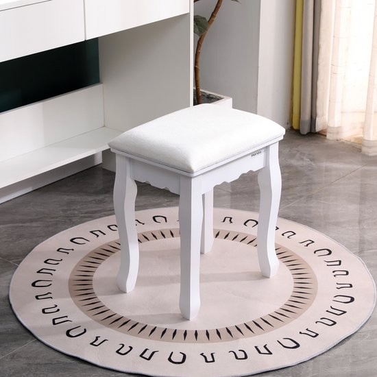 Witte gestoffeerde kruk op een ronde, crèmekleurige vloerkleed met zwarte cirkelvormige patronen. De kruk staat in een slaapkamer met een witte kast en plant op de achtergrond.