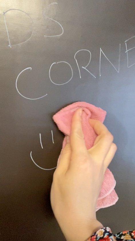Hand met roze doek veegt tekst 'DS CORNER' en een glimlach weg van een zwart magnetisch krijtbord.