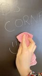 Hand met roze doek veegt tekst 'DS CORNER' en een glimlach weg van een zwart magnetisch krijtbord.