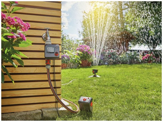Gardena besproeiingscomputer, aangesloten op een kraan, met sproeiende sprinkler in de tuin.