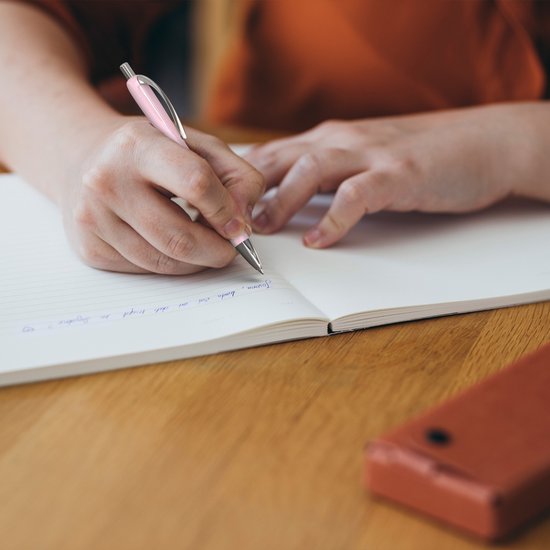 Een persoon schrijft in een schrift met een lichtroze Schneider Slider balpen.
