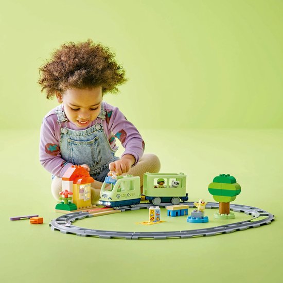 Un jeune enfant joue avec un train et des rails LEGO Duplo vert clair, avec une maisonnette et un arbre.