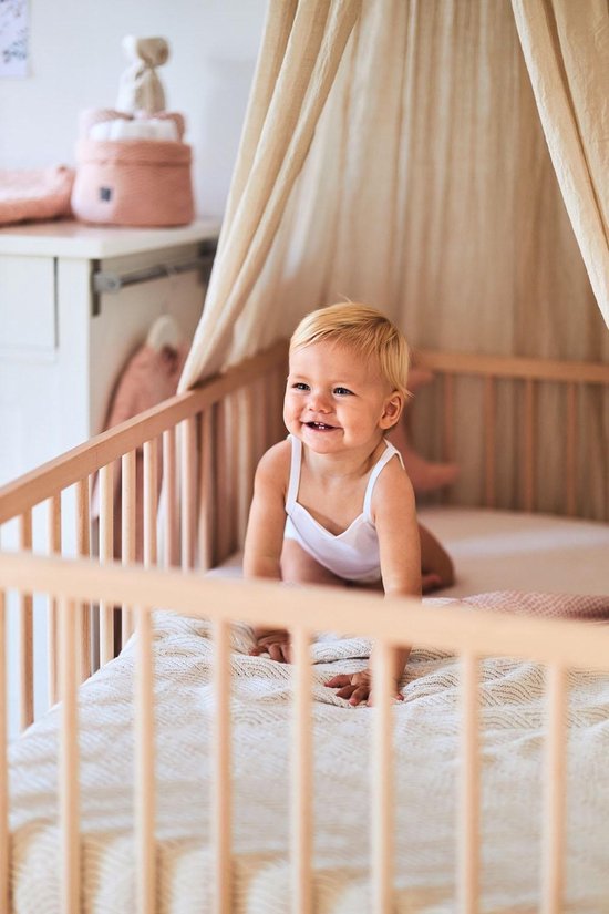 Een lachende baby in een wit hemdje kruipt in een ledikant onder een lichtbeige hemeltje.