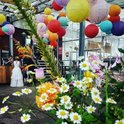 Een bruid in een witte jurk staat bij een bloemstuk, veelkleurige lampionnen erboven.
